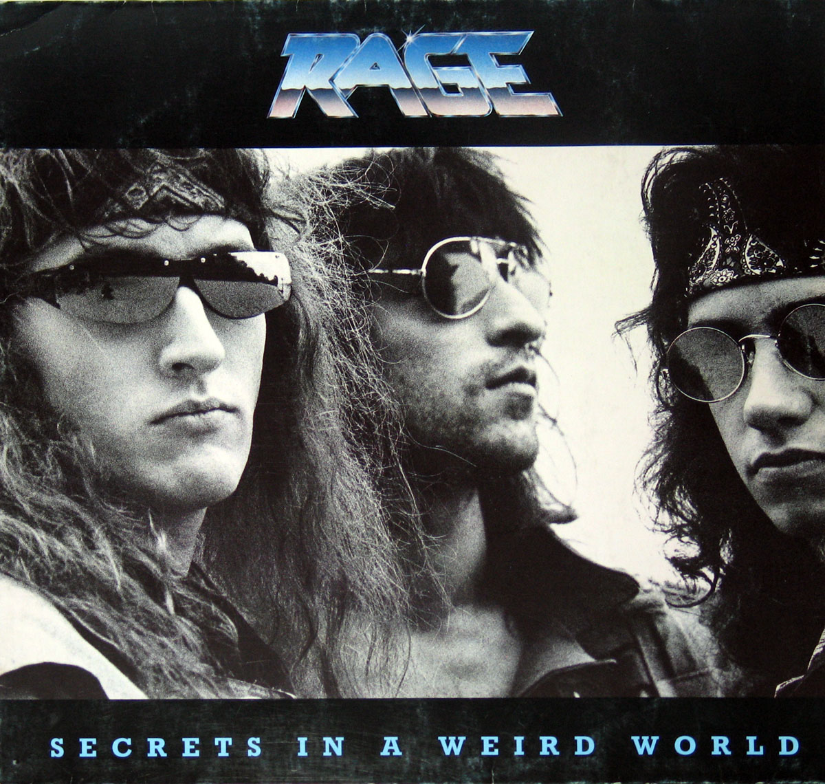 Album cover for Rage’s Secrets in a Weird World, featuring a black-and-white close-up of the three band members wearing sunglasses, with long hair and a classic 1980s metal look. The band logo is displayed at the top in metallic lettering, and the album title appears at the bottom in light blue text.